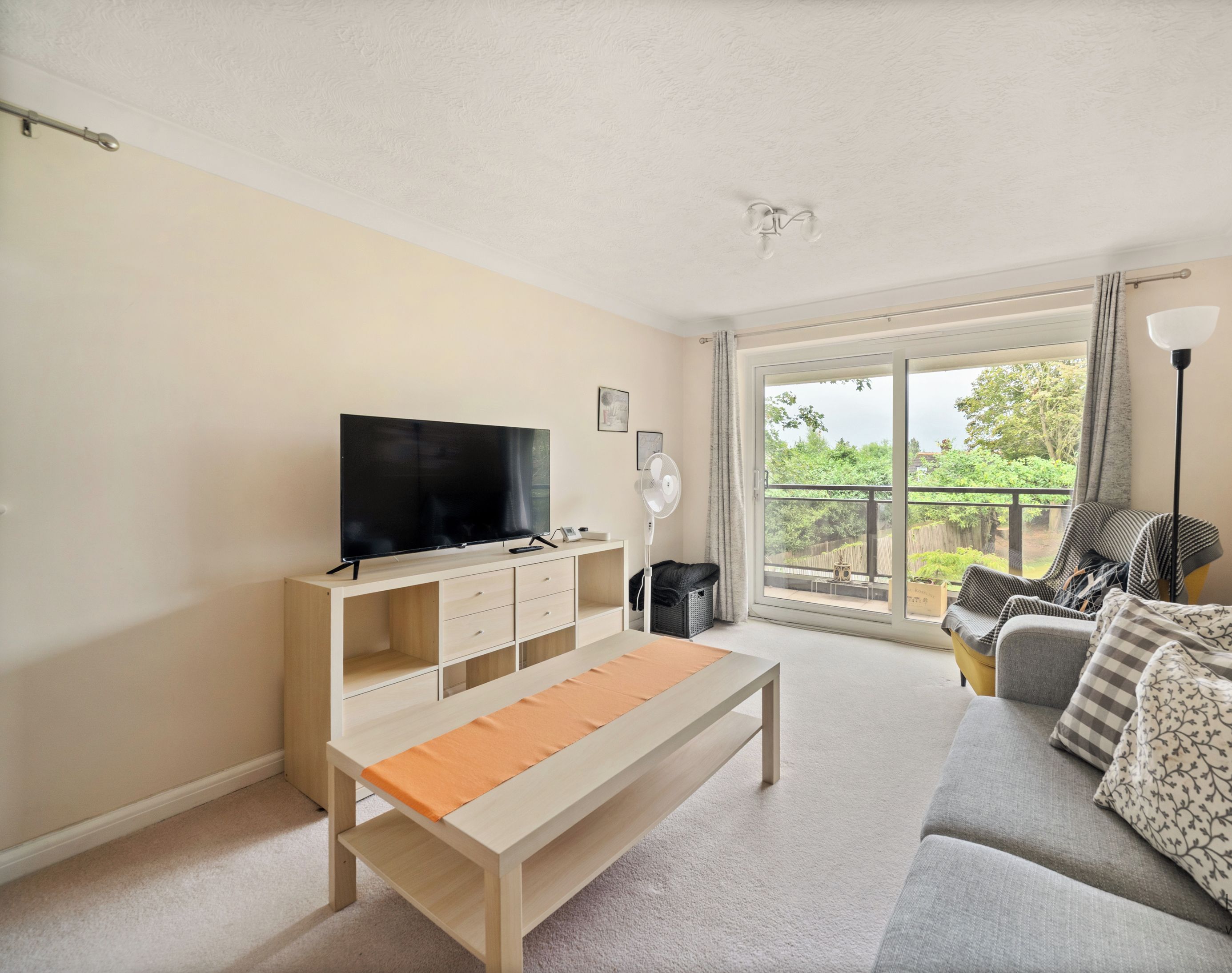 A living room with a TV, sofa, and a balcony with a view of greenery.