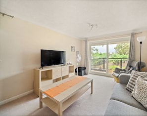 A living room with a TV, sofa, and a balcony with a view of greenery. - Walk to Bromley South Station & High Street, Free Parking, Sleeps 4 (Bromley)