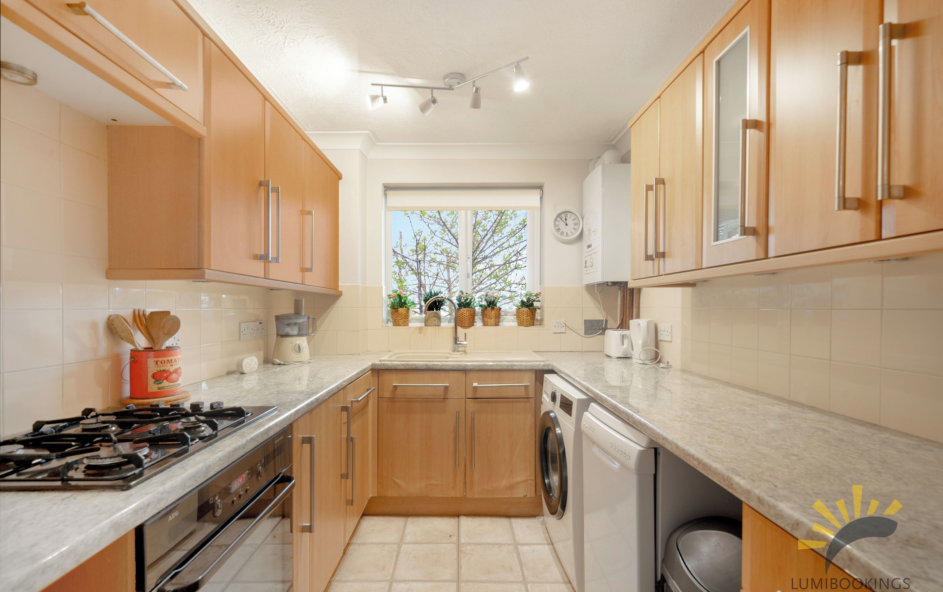 A modern kitchen with wooden cabinets, a gas stove, a washing machine, and a window with plants.