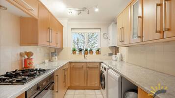 A modern kitchen with wooden cabinets, a gas stove, a washing machine, and a window with plants.