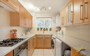 A modern kitchen with wooden cabinets, a gas stove, a washing machine, and a window with plants. - Walk to Bromley South Station & High Street, Free Parking, Sleeps 4 (Bromley)
