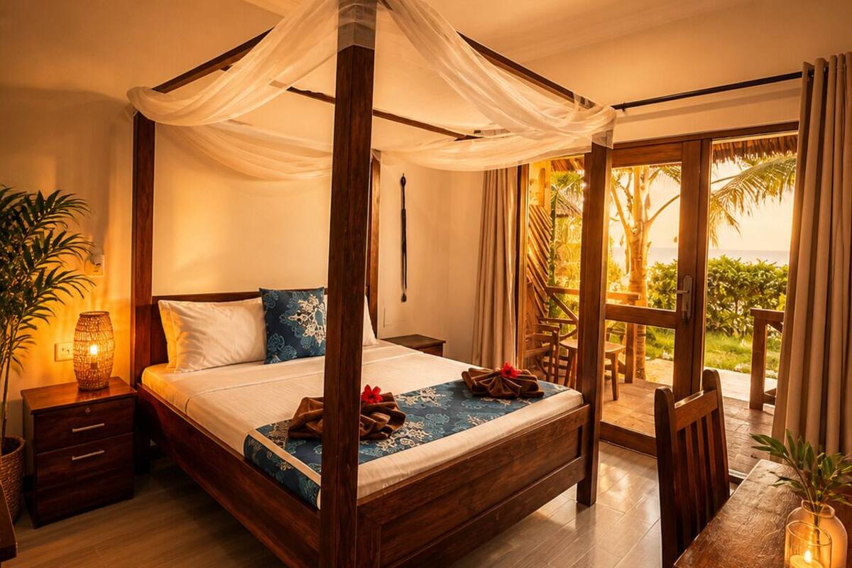 A four-poster bed with a canopy, a dining table with chairs, a small table with a candle, and a view of palm trees and the ocean.