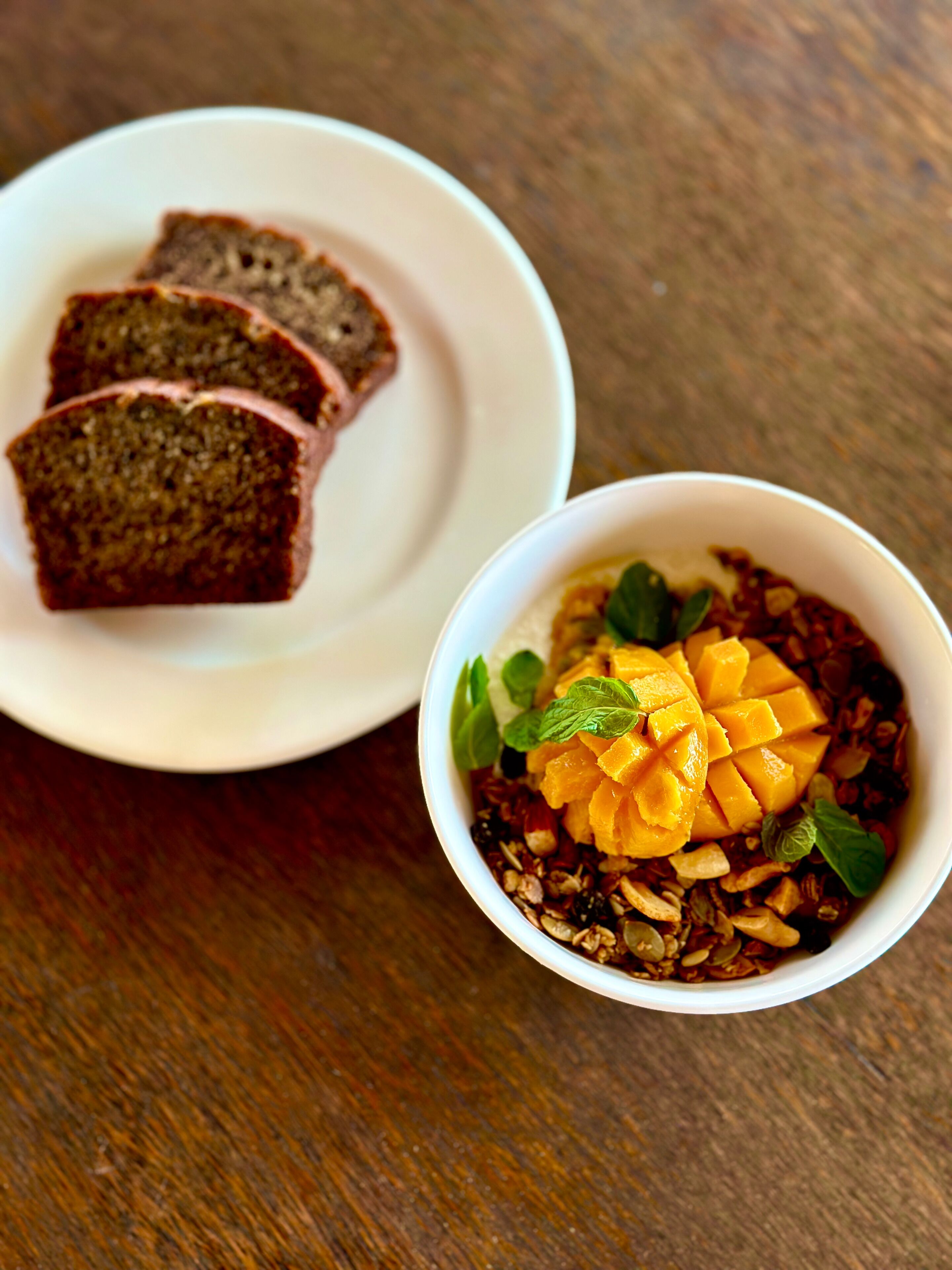 A bowl of granola with mango chunks and mint leaves, and a plate with slices of dark bread.