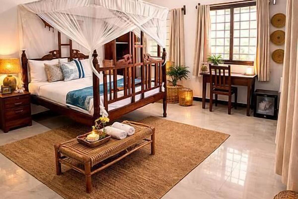 A bedroom with a canopy bed, a ceiling fan, a bedside table, a chair, a small table, and a window with curtains.