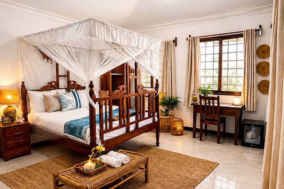 A bedroom with a four-poster bed, a ceiling fan, a bedside table, a nightstand, a bedside lamp, a window with curtains, a small table with a lamp, and a wicker chair.