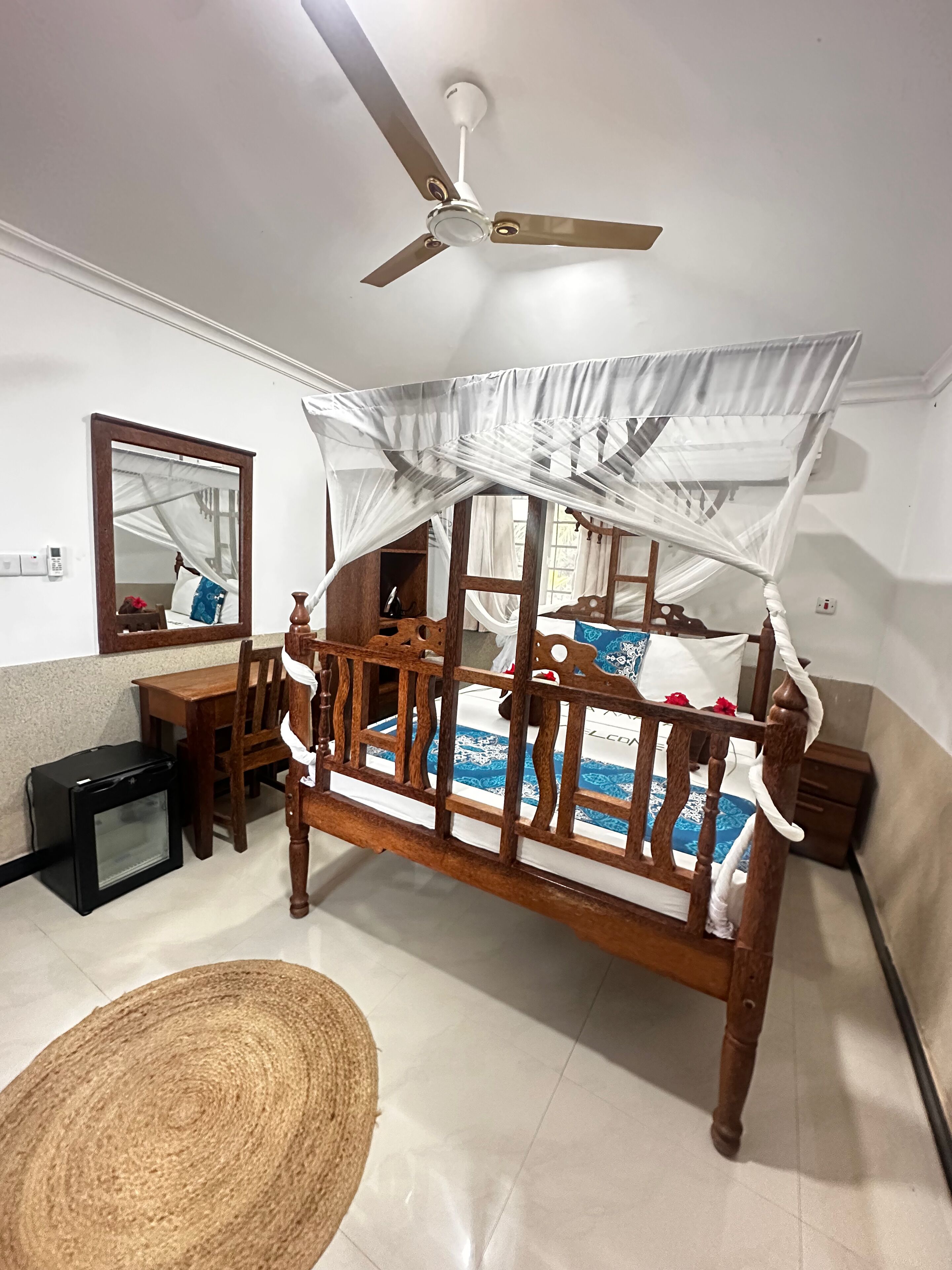 Une chambre à coucher avec un lit à baldaquin en bois, un ventilateur de plafond, une table de chevet, un petit bureau, une chaise et un tapis tissé.