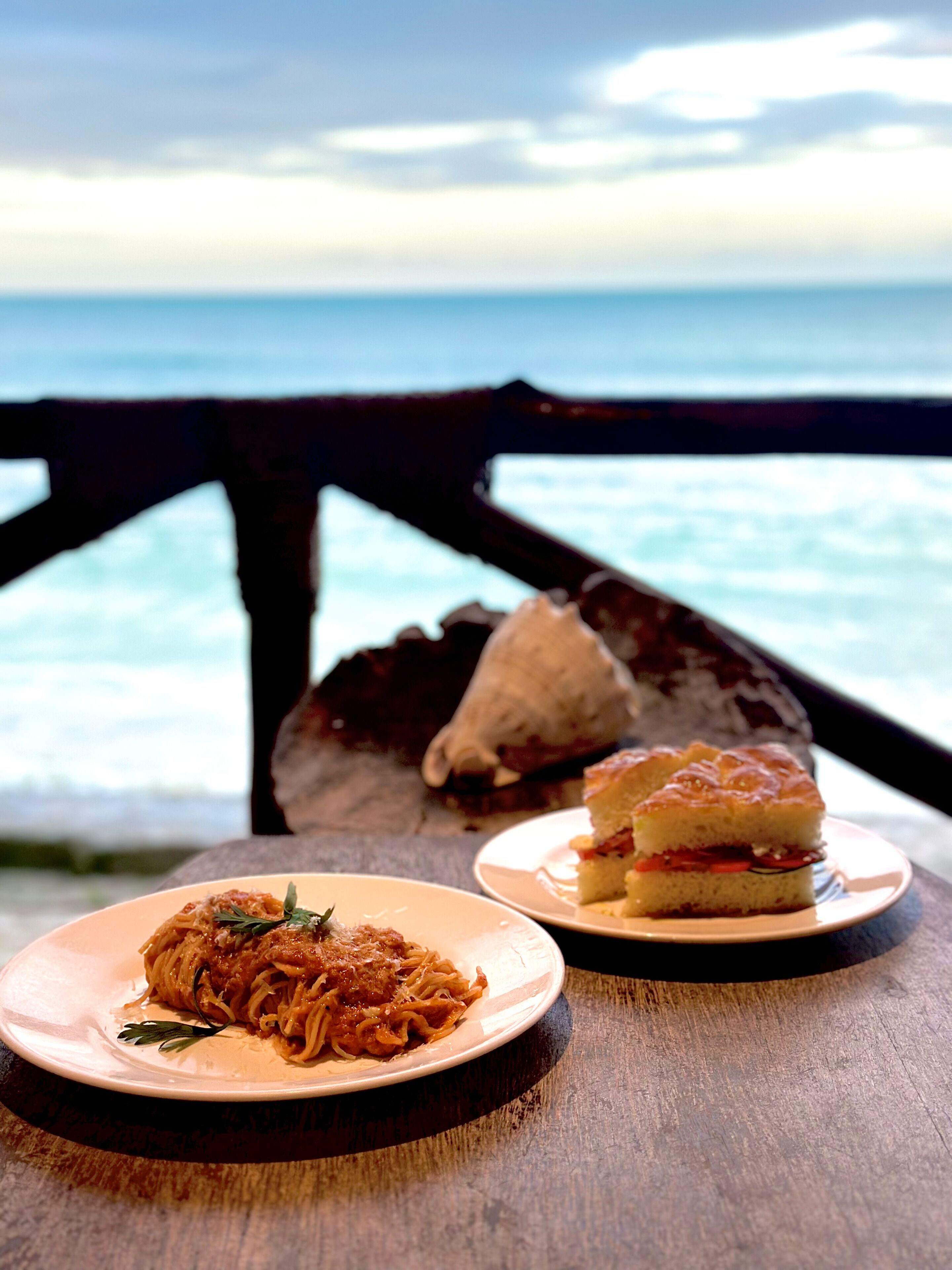 Un tavolo con due piatti di cibo: uno con pasta e l'altro con panini, sullo sfondo un binario in legno con vista su un corpo d'acqua.