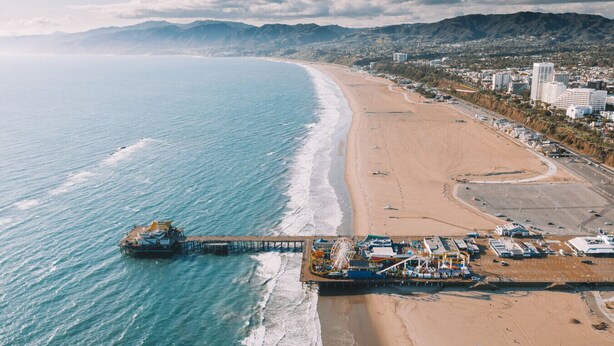 An aerial view of a beach with a pier extending into the ocean, a sandy beach, and a cityscape in the background.