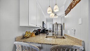 A kitchen with white cabinets, a granite countertop, a sink, and three hanging pendant lights. - Coco Cabana - 4th Floor w/ Indoor & Outdoor Pools! (Hilton Head)