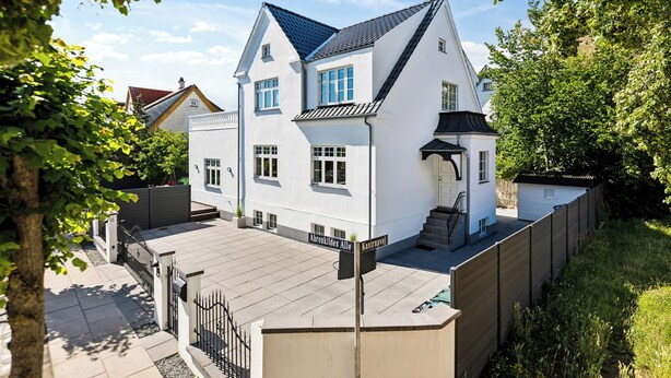 Ein zweistöckiges weißes Haus mit dunklem Dach, einer kleinen angebauten Garage und einem gepflasterten Einfahrt.