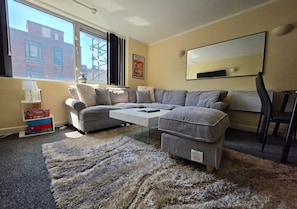 A living room with a sectional sofa, a coffee table, a rug, a TV, and a window with a view of buildings. - Stylish 1BR Flat Next to Hockley (Nottingham)