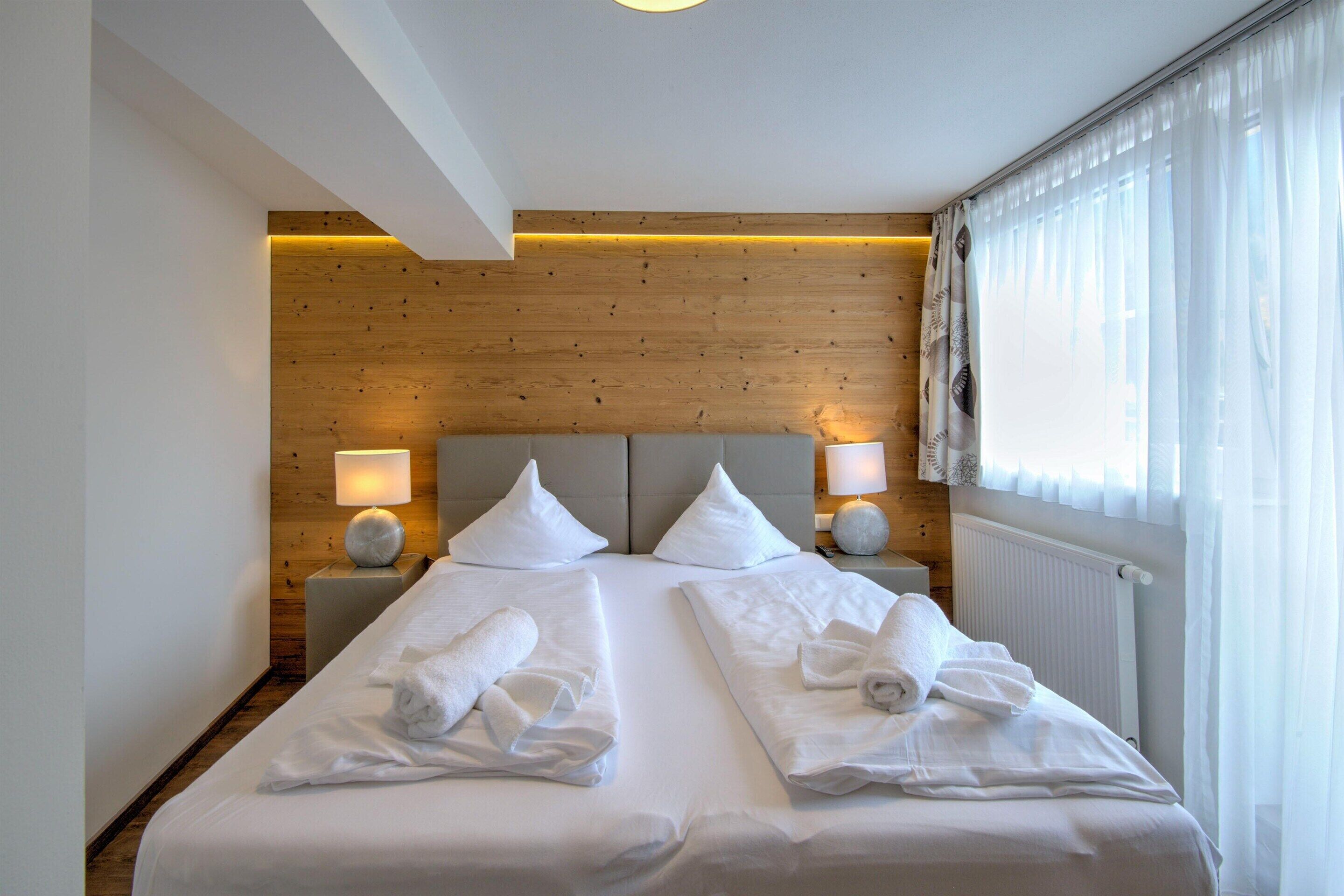 A double bed with white linens and towels, wooden panel walls, two bedside lamps, and a window with sheer curtains.