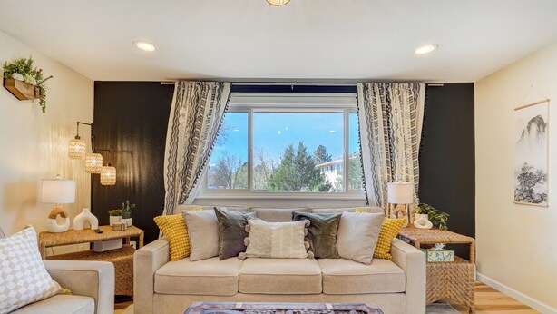 A living room with a beige sofa, patterned cushions, a wooden coffee table with a carved design, and a large window with curtains.