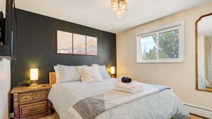 A bedroom with a bed, bedside lamps, a dresser, a mirror, and a window with a view of trees.