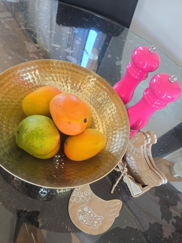 A glass table with a decorative bowl containing fruits, a wooden bird-shaped salt shaker, and two pink pepper mills.