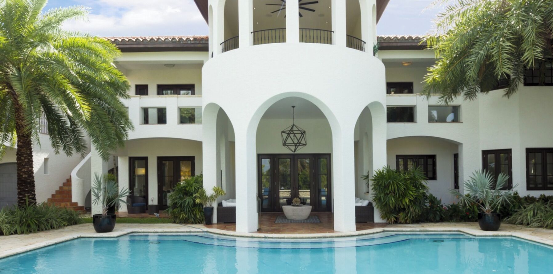 A poolside view of a two-story building with a covered patio area, outdoor seating, and a large planter.
