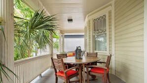 Een overdekte veranda met een houten tafel en stoelen, omringd door witte leuningen en een grote potplant.