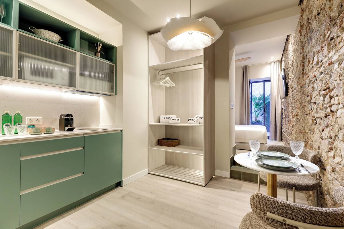 Eine moderne Küche mit grünen Schränken, ein Essbereich mit Tisch für zwei Personen und ein Blick ins Freie durch ein Fenster.