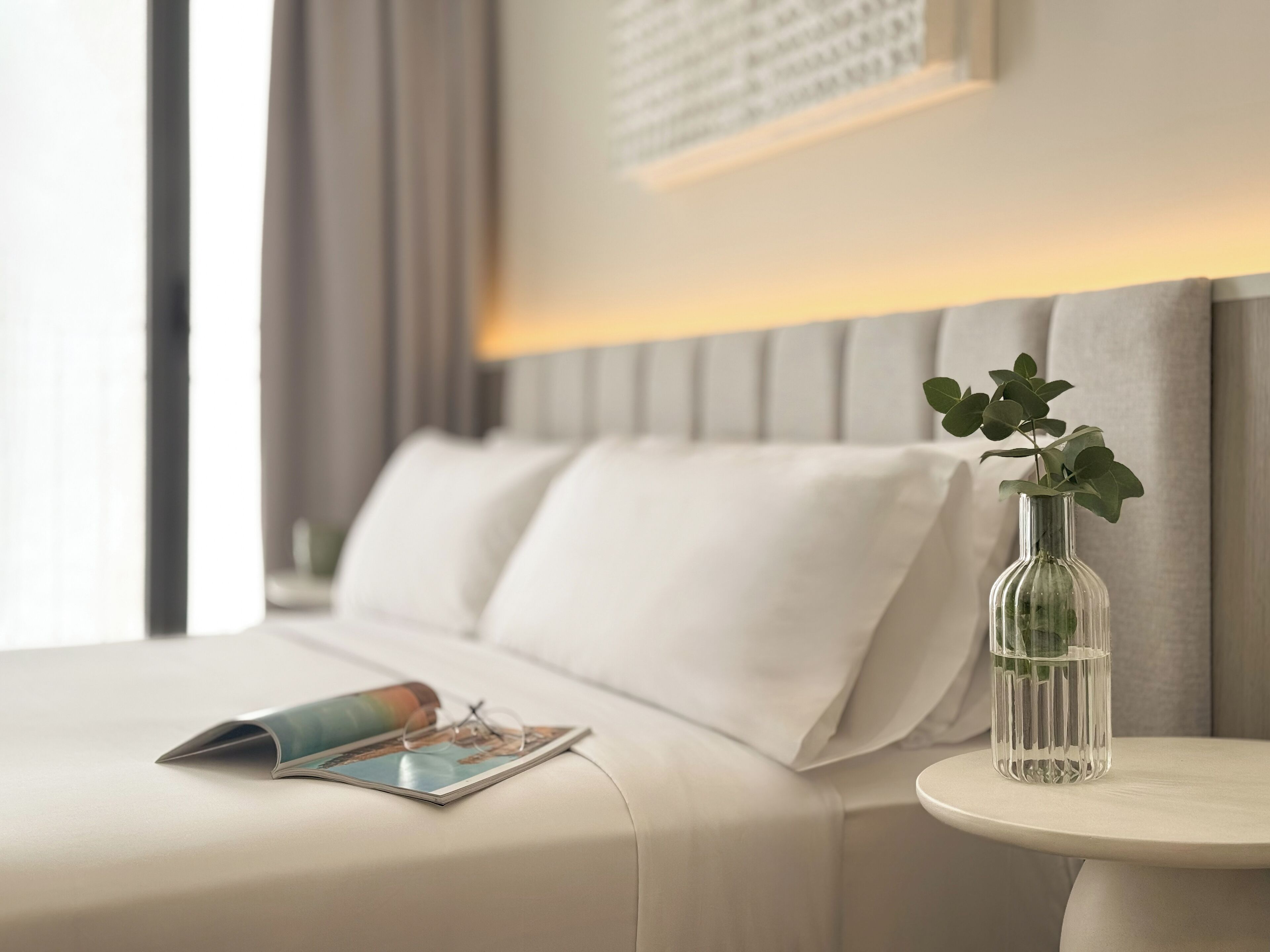 A neatly made bed with white linens, a bedside table with a clear vase containing green leaves, and a magazine on the bed.