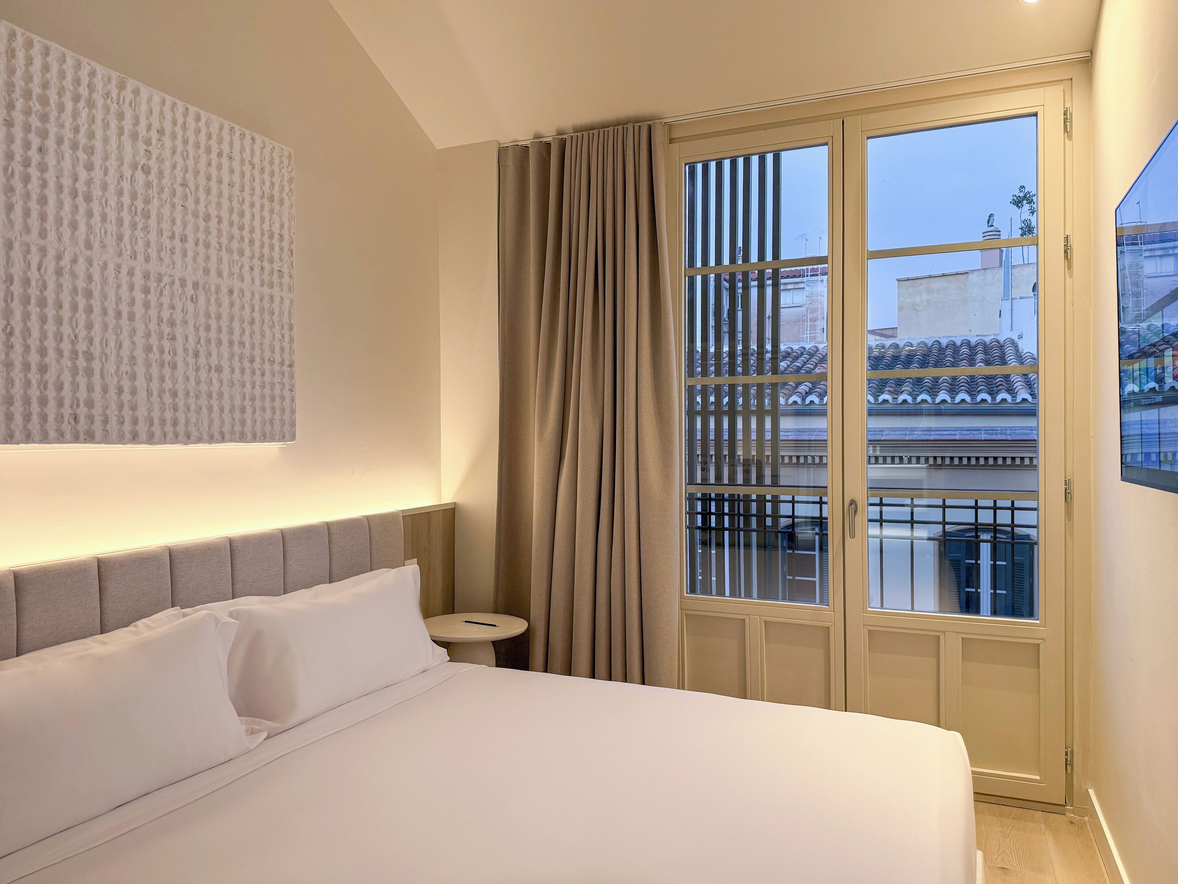 A hotel room with a bed, a small bedside table, a window with curtains, and a view of the outside.