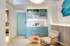 Una cocina compacta con armarios verde azulado, una silla de madera y una pequeña mesa con fruta y vasos.