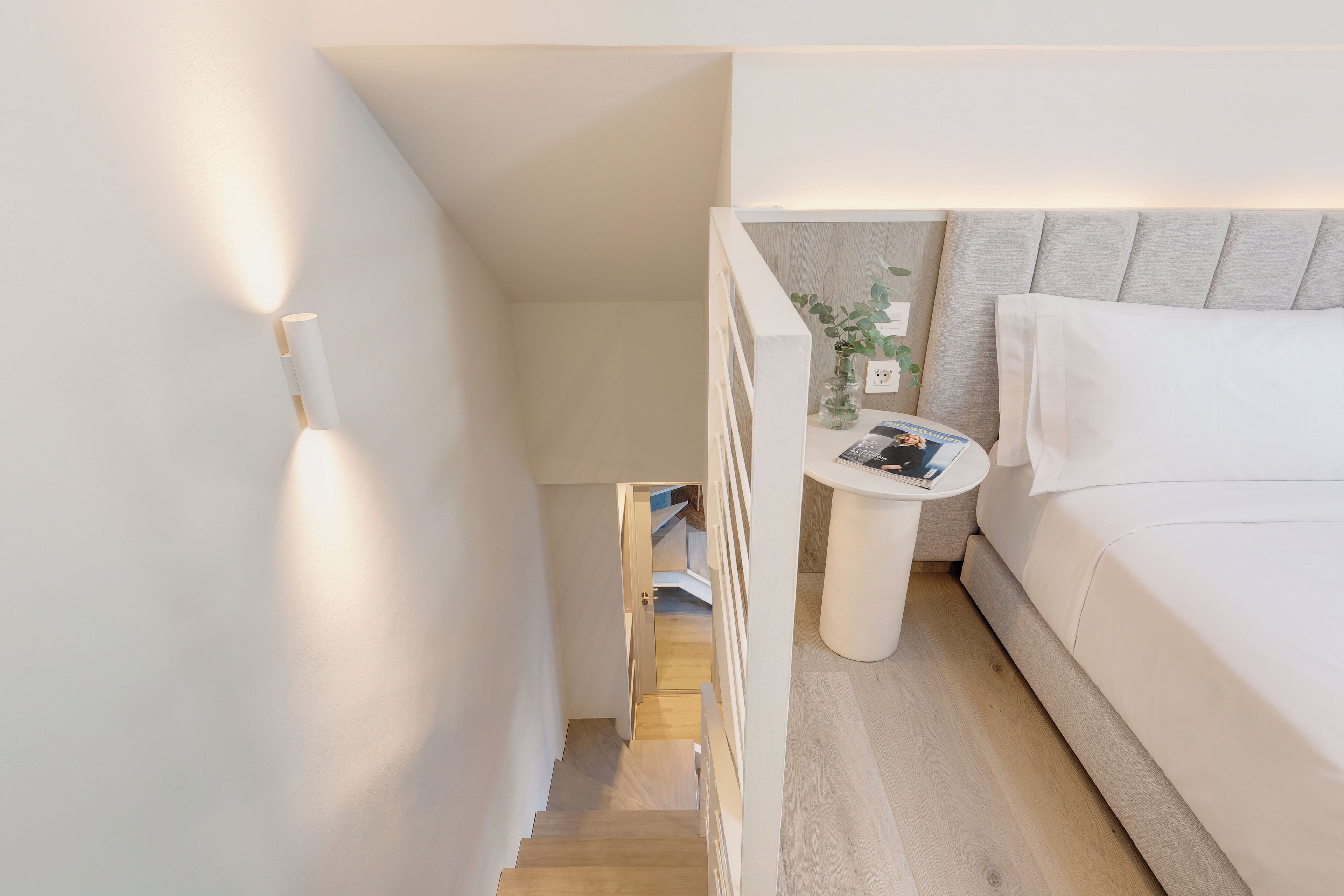 Et moderne soveværelse med en hvid seng, et natbord med en lampe, og udsigt til trappen.