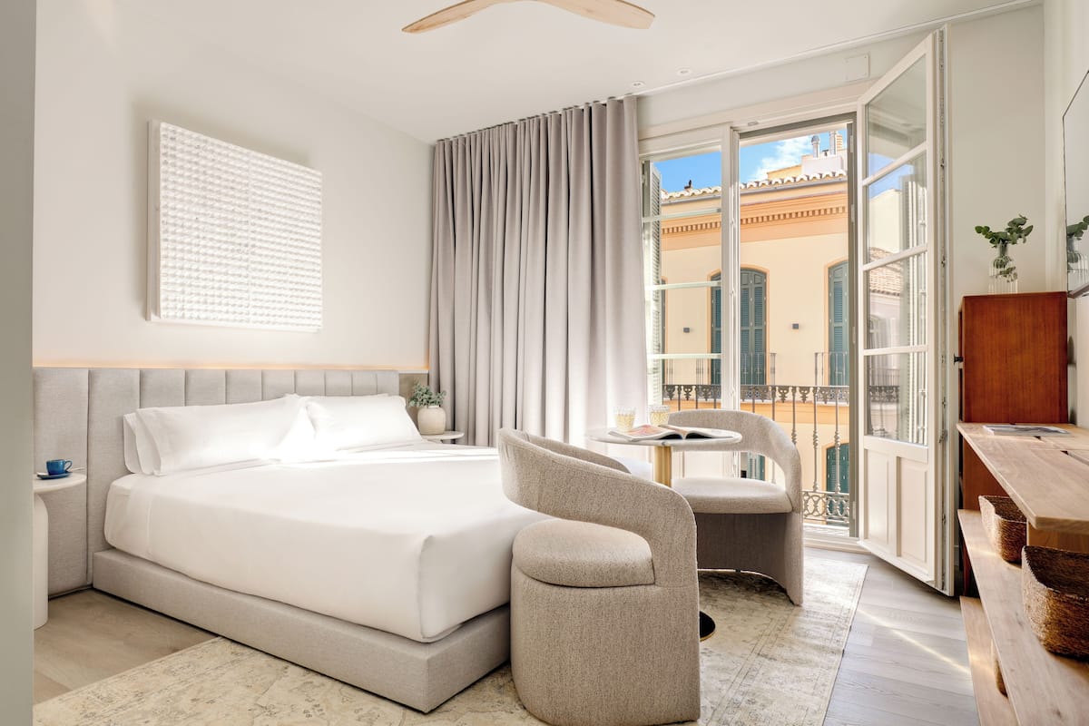 Ein Hotelzimmer mit Bett, Stuhl, Schreibtisch und einem Balkon mit Blick auf Gebäude.