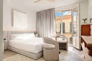 Habitación de hotel con cama, una silla, escritorio y balcón con vistas a edificios.