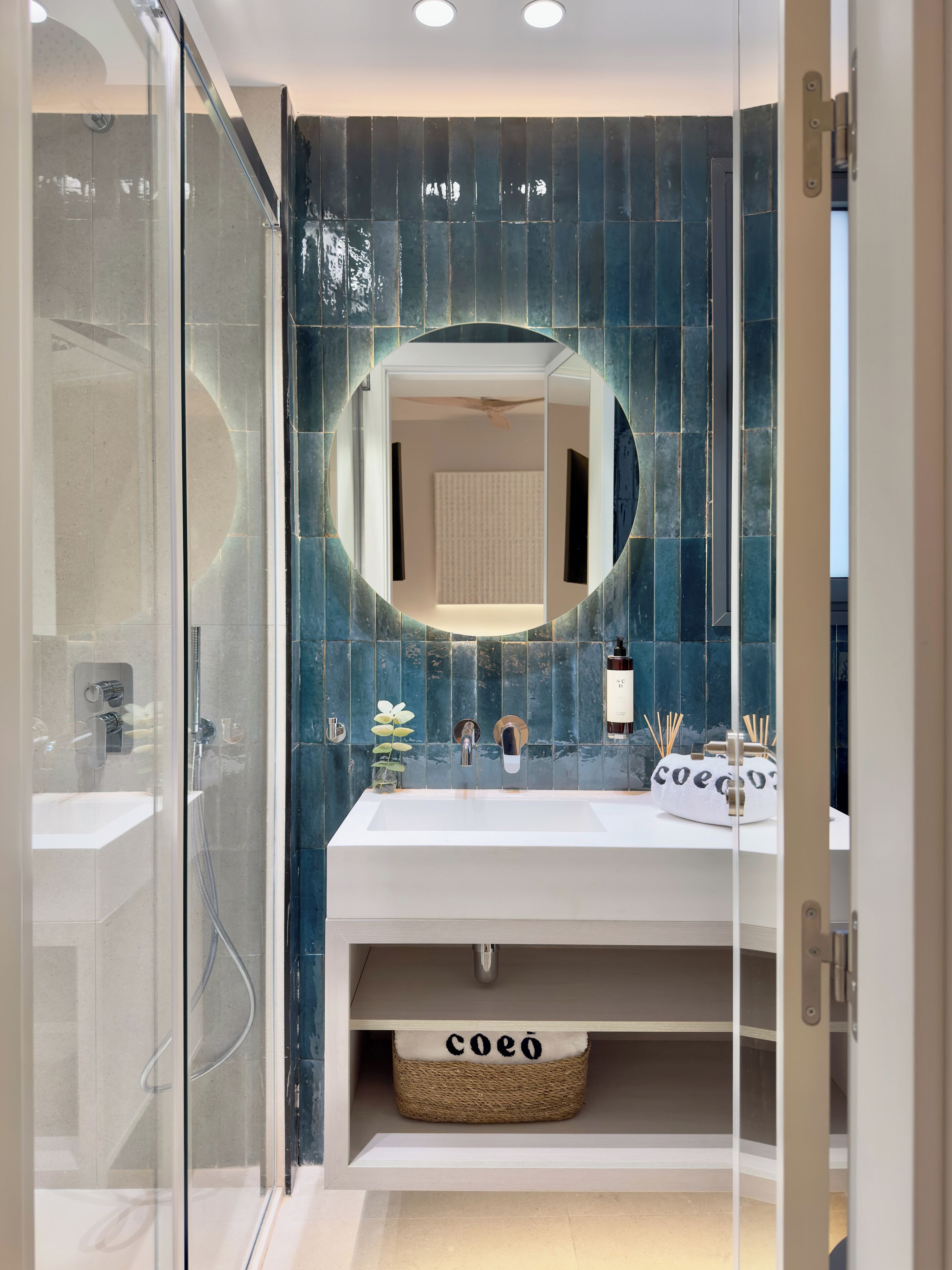 Et moderne badeværelse med en glasbrusekabine, et hvidt toiletbord med et rundt spejl og en væg med blå fliser.