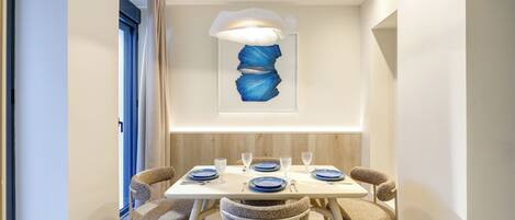 A dining area with a white table set for four, featuring blue plates and clear glassware, a modern abstract painting on the wall, and a unique ceiling light fixture.