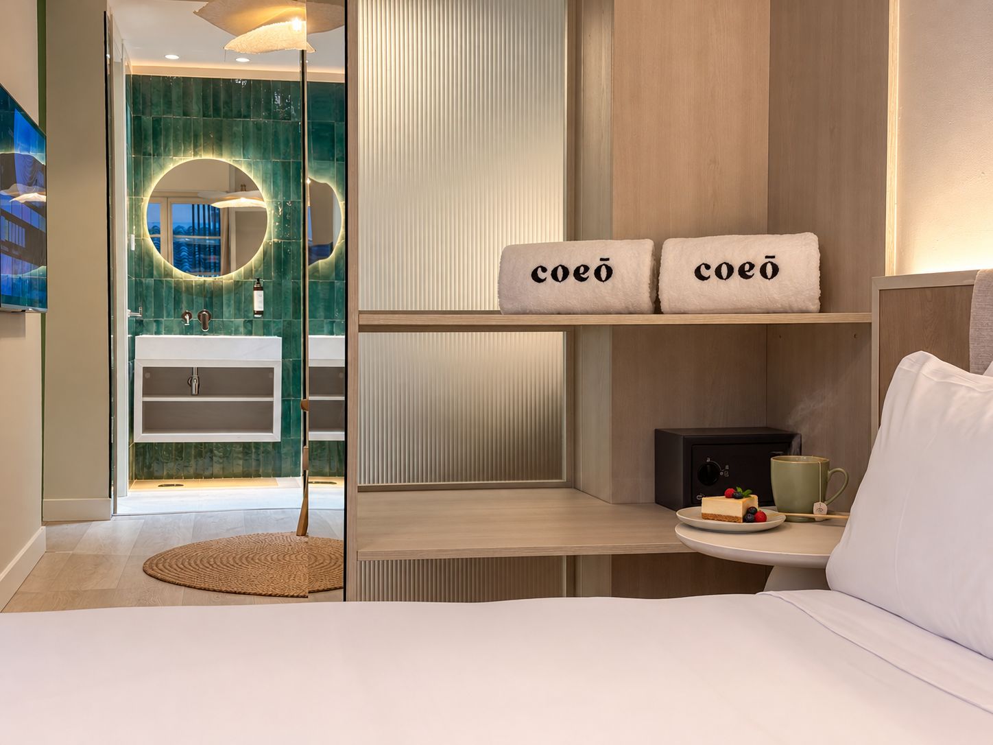 A modern bedroom with a bed, a bedside table with a cup and a plate of fruit, and a view of a bathroom with a glass shower.