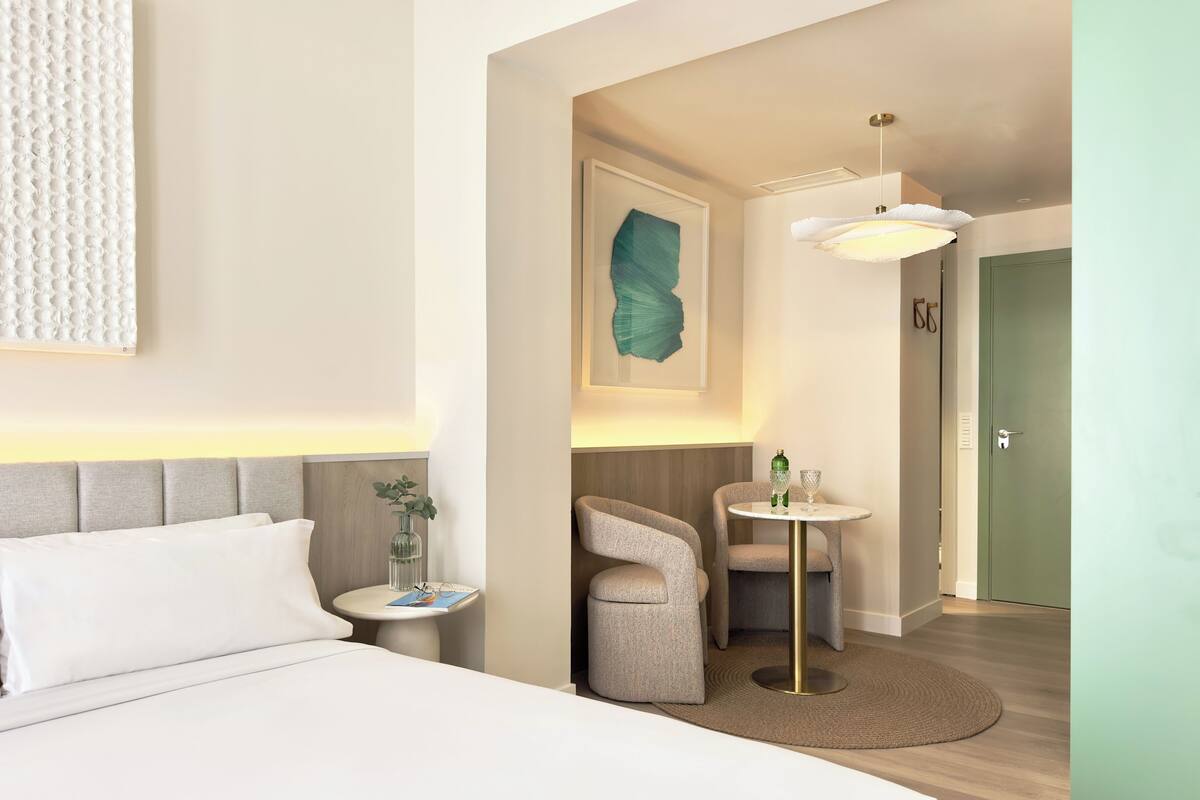 Ein Hotelzimmer mit einem Bett, einem kleinen runden Tisch mit zwei Stühlen, einem Nachttisch, einer grünen Vase und einer türkis farbenen Wandkunst.