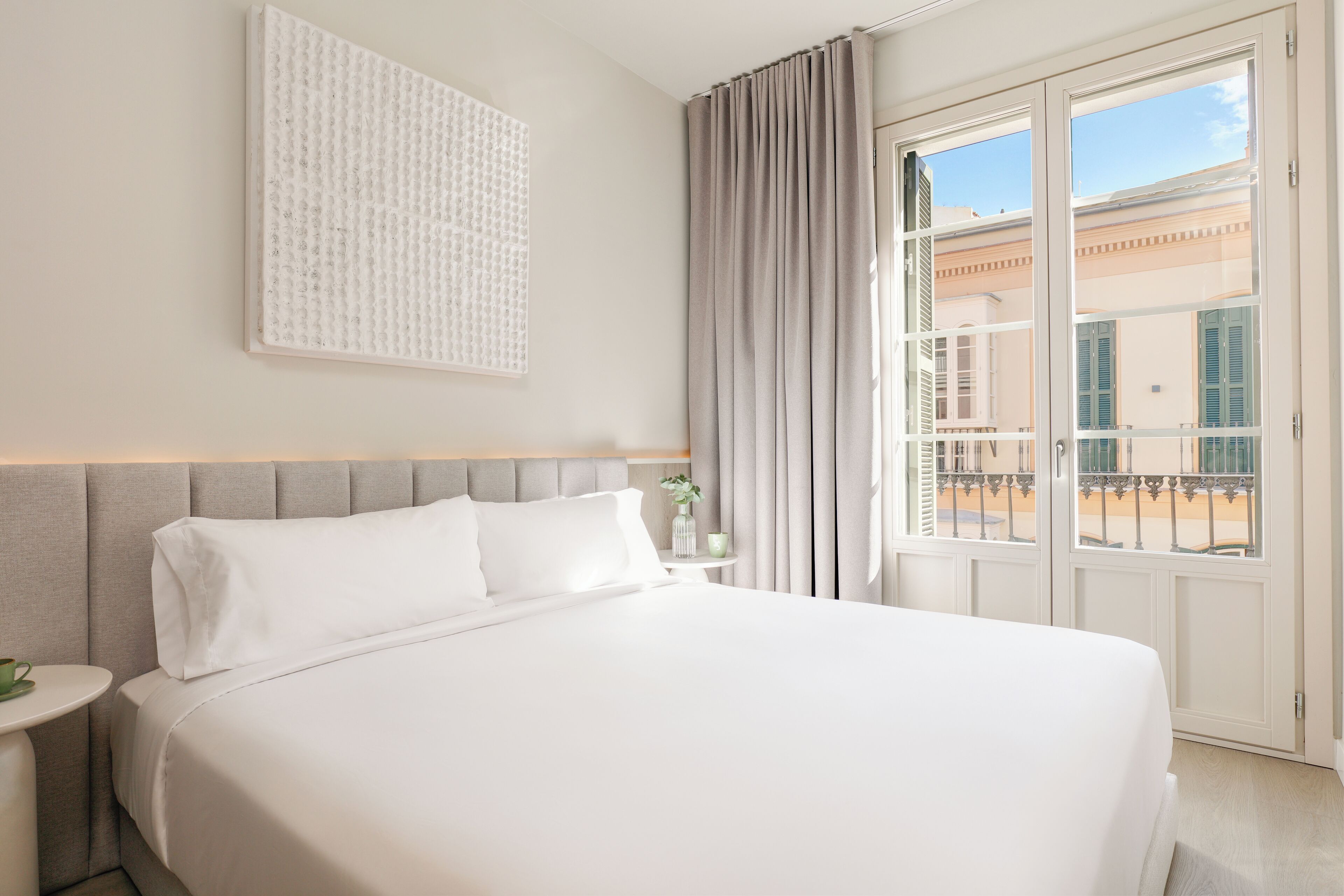 Una cama bien hecha con cabecera, una mesita de noche con una taza verde y una ventana con cortinas.