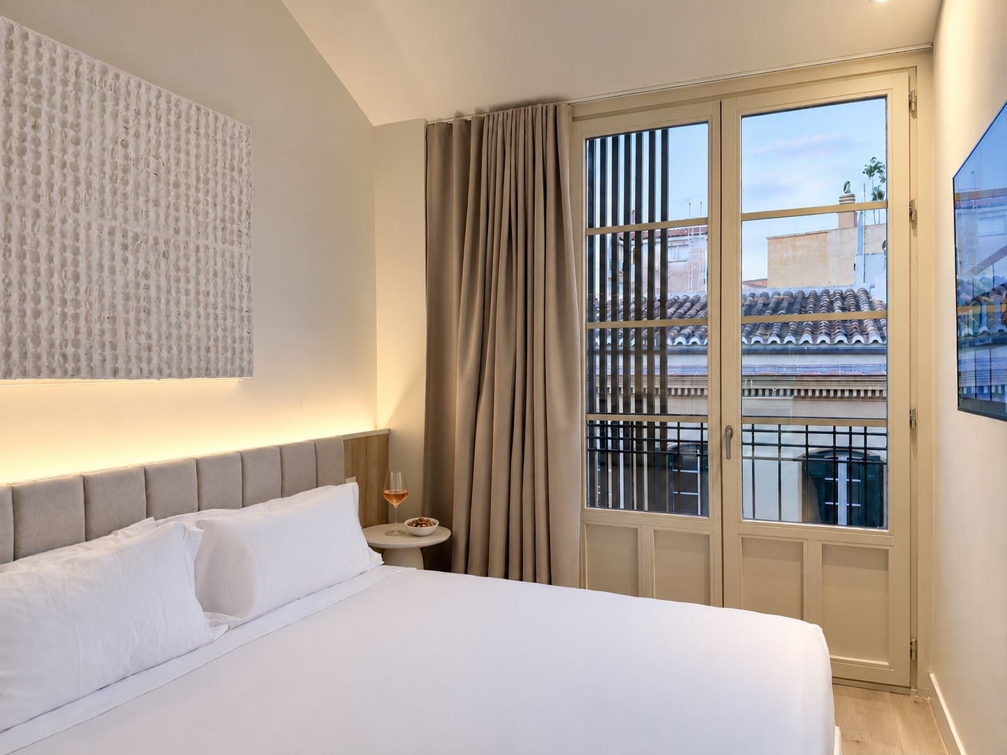 A hotel room with a bed, a small table with a glass of wine, and a view of a building through a window with curtains.
