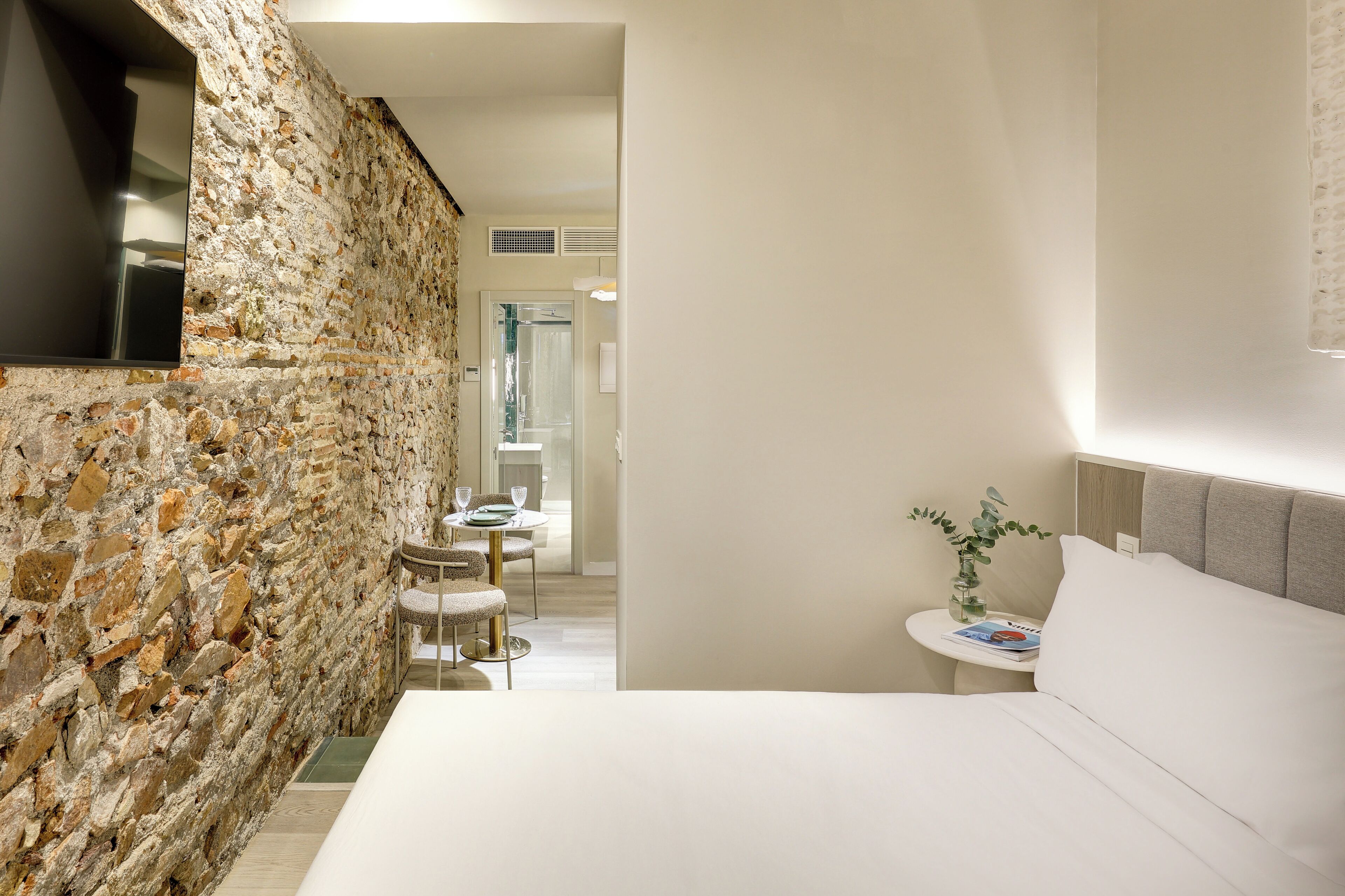 A modern hotel room with a stone accent wall, a bed with white linens, a small round table with a vase, and a view into the dining area.