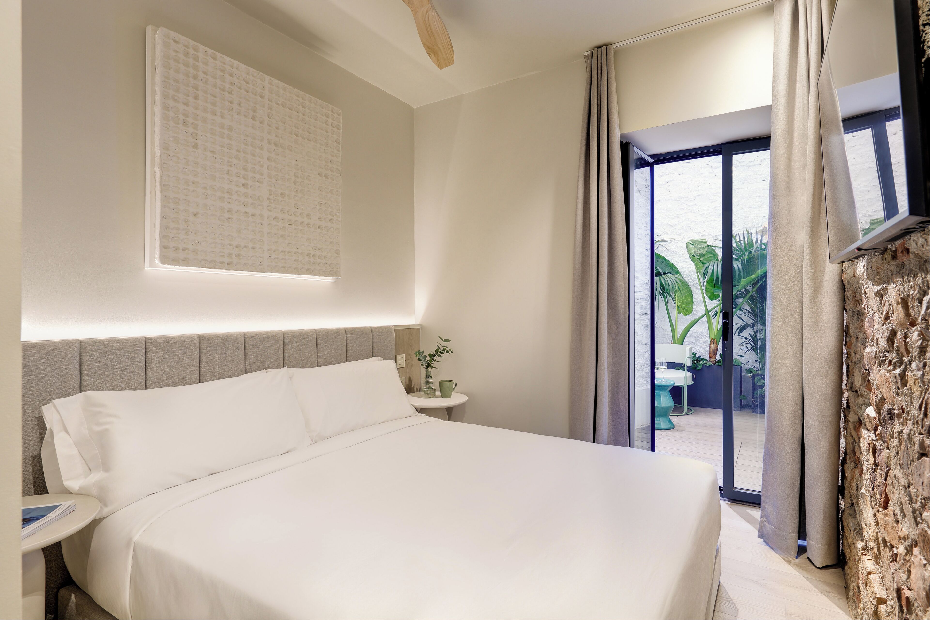 A hotel room with a large bed, a small bedside table with a lamp, a wall-mounted TV, and a view of the outdoors through a glass door.