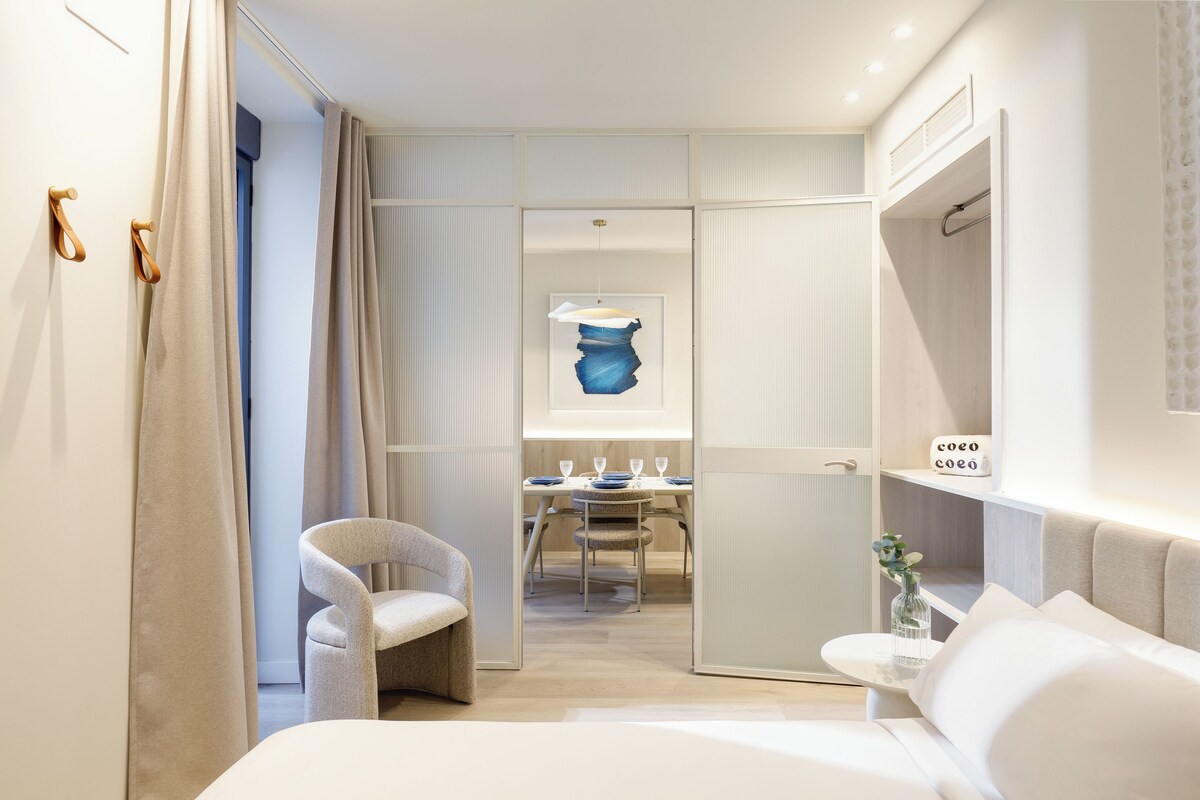 A modern hotel room with a bed, a chair, a dining table with chairs, a small table with a vase, and a wardrobe.