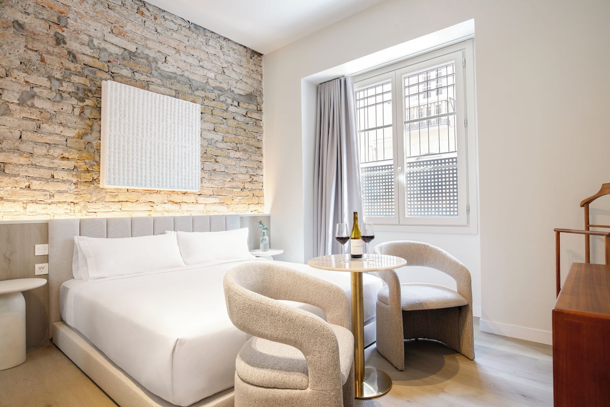Ein modernes Hotelzimmer mit Backsteinwand, einem großen Bett, zwei Sesseln, einem kleinen Tisch mit einer Weinflasche und einem Fenster mit Vorhängen.