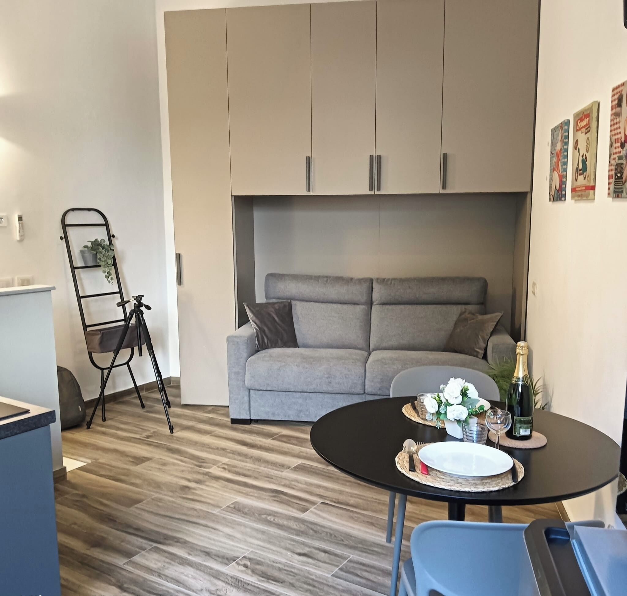 Un espace de vie compact avec un canapé gris, une petite table ronde sur laquelle se trouve une bouteille de champagne, et une échelle en métal noir adossée au mur.