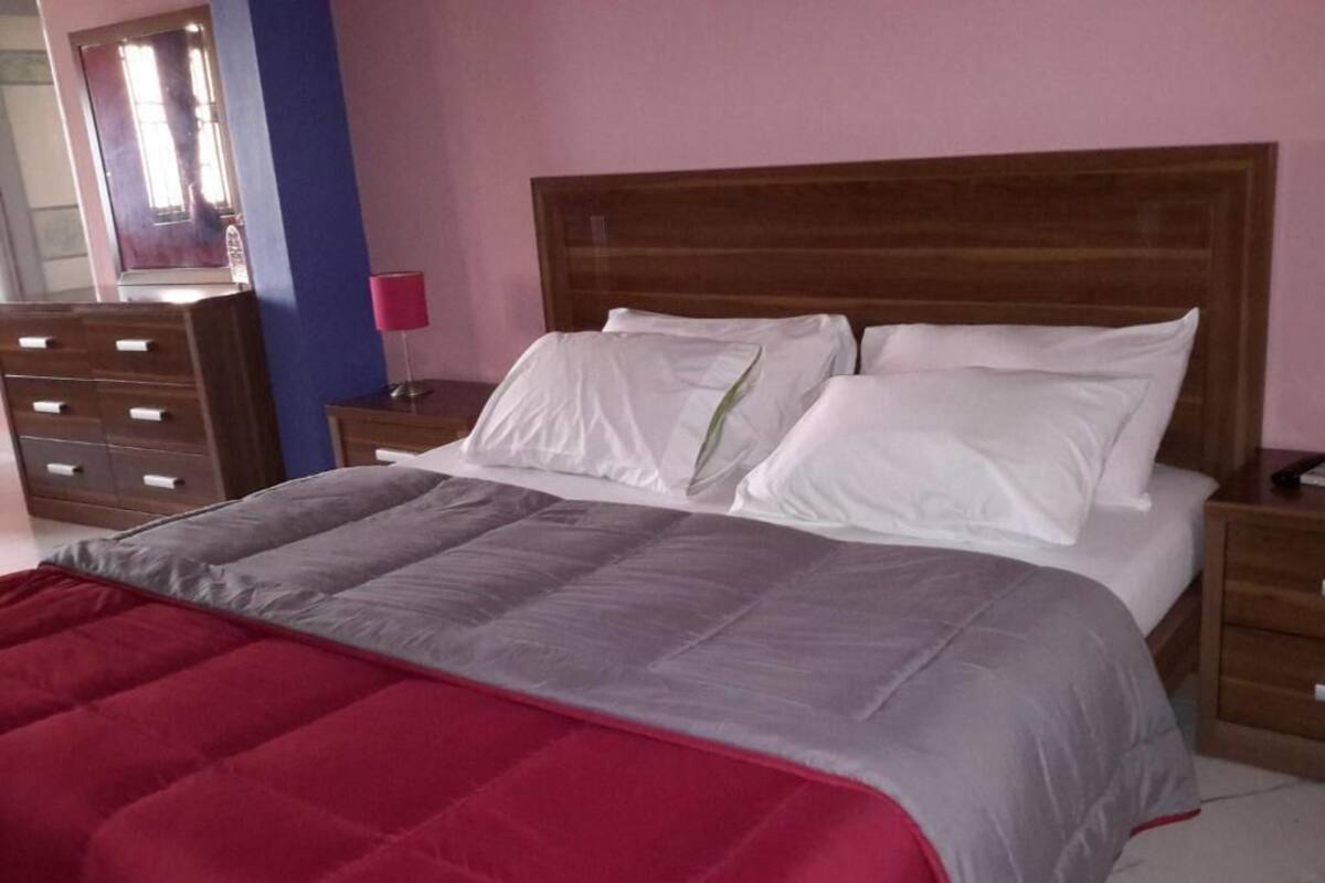 A bedroom with a wooden headboard, a bed with white bedding, a red and grey blanket, a wooden dresser, and a nightstand with a lamp.