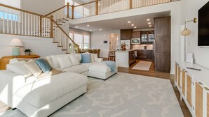 A spacious living area with a white sectional sofa, a staircase leading to an upper level, and a kitchen area with dark wooden cabinets. - Resort Retreat: King Suites & Nature Trails (Millsboro)
