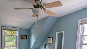 Un dormitorio con techo inclinado, ventilador de techo, escritorio, silla, cama y dos ventanas.