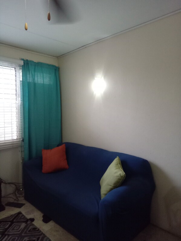 A room with a blue sofa, a red and a green pillow, a window with blinds, and a light fixture on the wall.