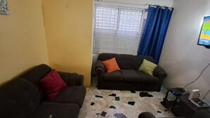 Una sala de estar con dos sofás, piso de baldosas, una ventana con persianas y un televisor en la pared.