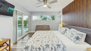 Une chambre avec un lit, un ventilateur de plafond, un téléviseur fixé au mur, une fenêtre donnant sur un espace verdoyant et une porte-fenêtre coulissante qui mène à un espace extérieur.