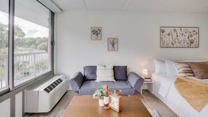 Una camera d'albergo moderna con un divano grigio, un letto bianco e un tavolino in legno con una pianta e una sveglia.