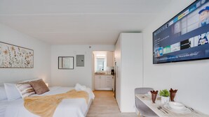 Una moderna camera d'albergo con un letto, una scrivania con una sedia, una TV a parete e un angolo cottura sullo sfondo.