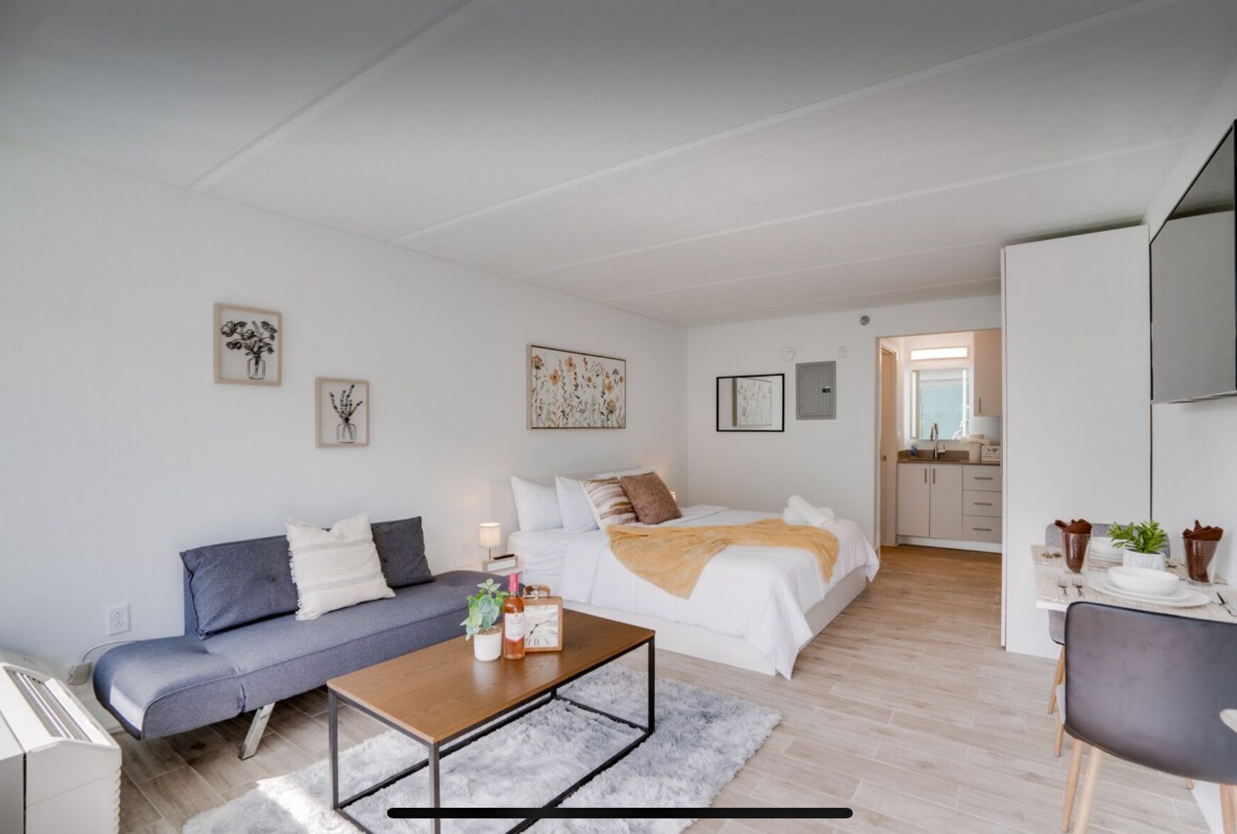 A modern, well-lit apartment with a large bed, a sofa, a dining table, and a kitchen area.