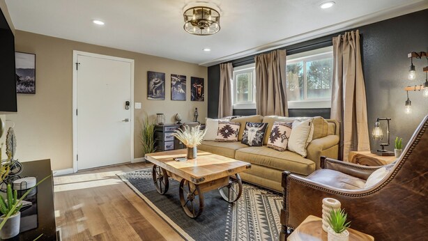 A cozy living room with a sofa, coffee table, and armchair, a dining table with chairs, and a large window with curtains.