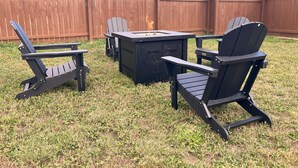 An outdoor seating area with Adirondack chairs and a square table, set against a wooden fence and a grassy lawn.